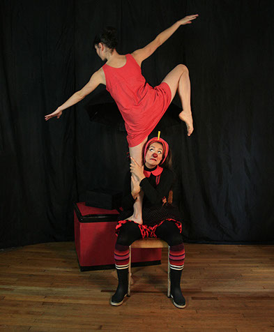 Le clown, assis sur une chaise, tient maladroitement la danseuse en train de faire une figure debout sur la chaise.