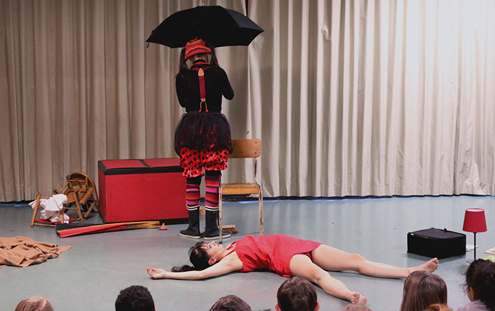 la dasneuse est par allongée par terre. Le clown est en train d'attacher un parapluie à une chaise.