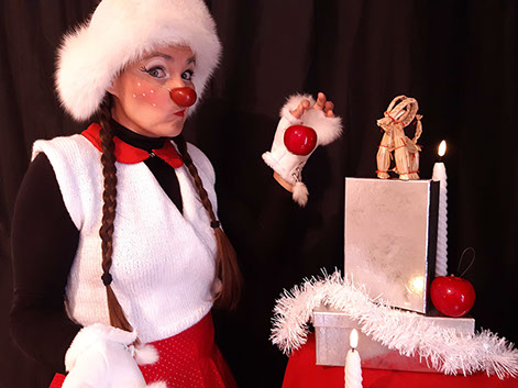 Photo. Le clown, Mlle Slurp, tente de mettre une boule de noël sur un sapin fait avec des boites.